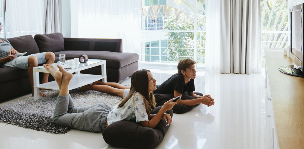 Vuelta al cole y a la rutinas. Imagen de un padre con una tableta en un sofá mientras sus hijos adolescentes (chico y chica) ven la televisión.