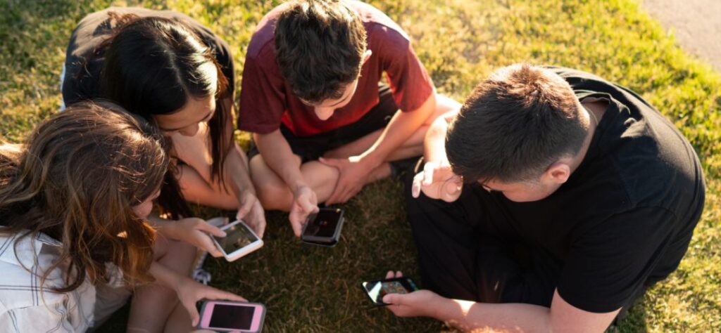 Plano cenital de 4 adolescentes sentados sobre la hierba, cada uno con su móvil